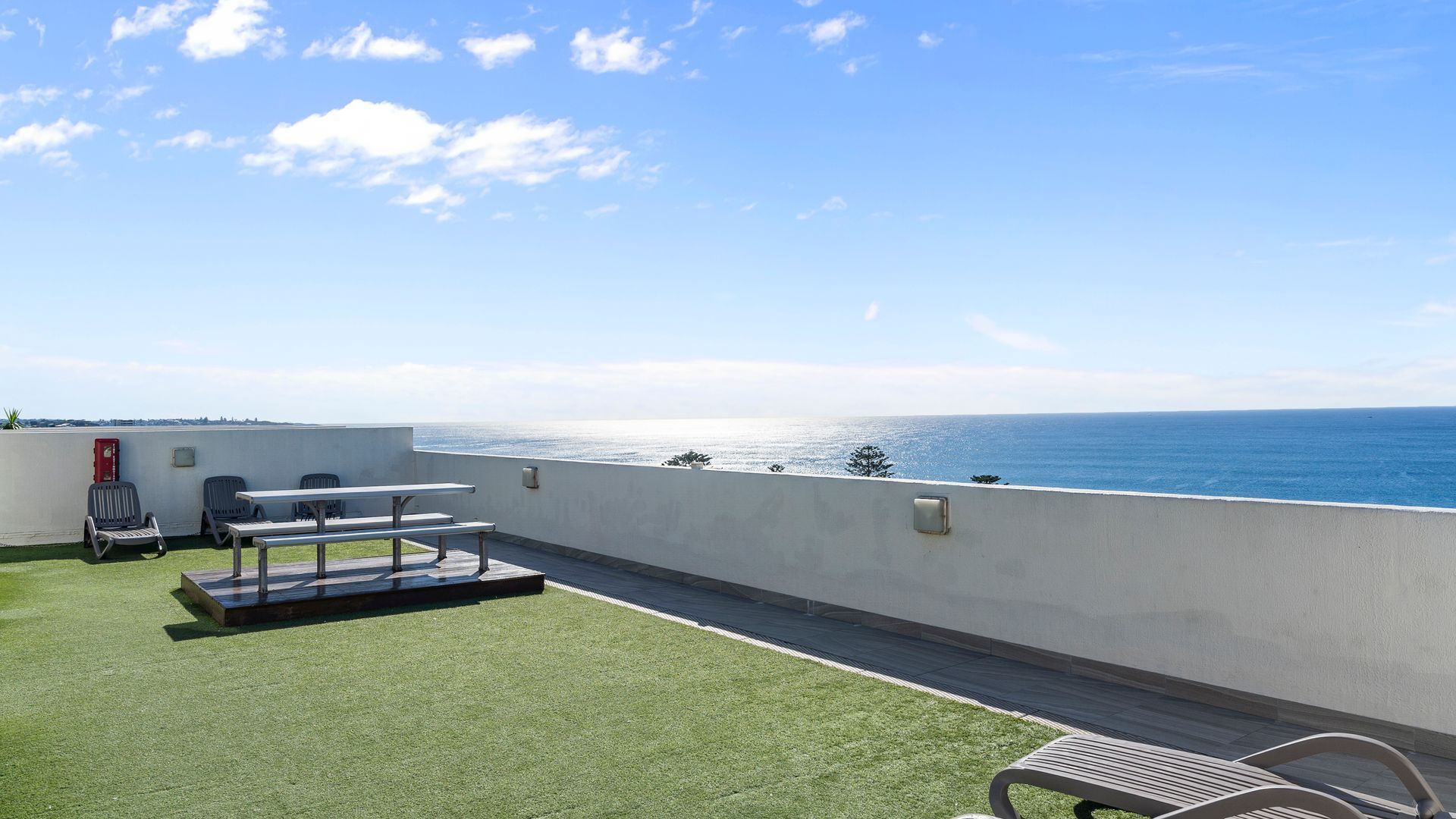 A rooftop terrace with a view of the sea, a table, chairs, and a small garden area.