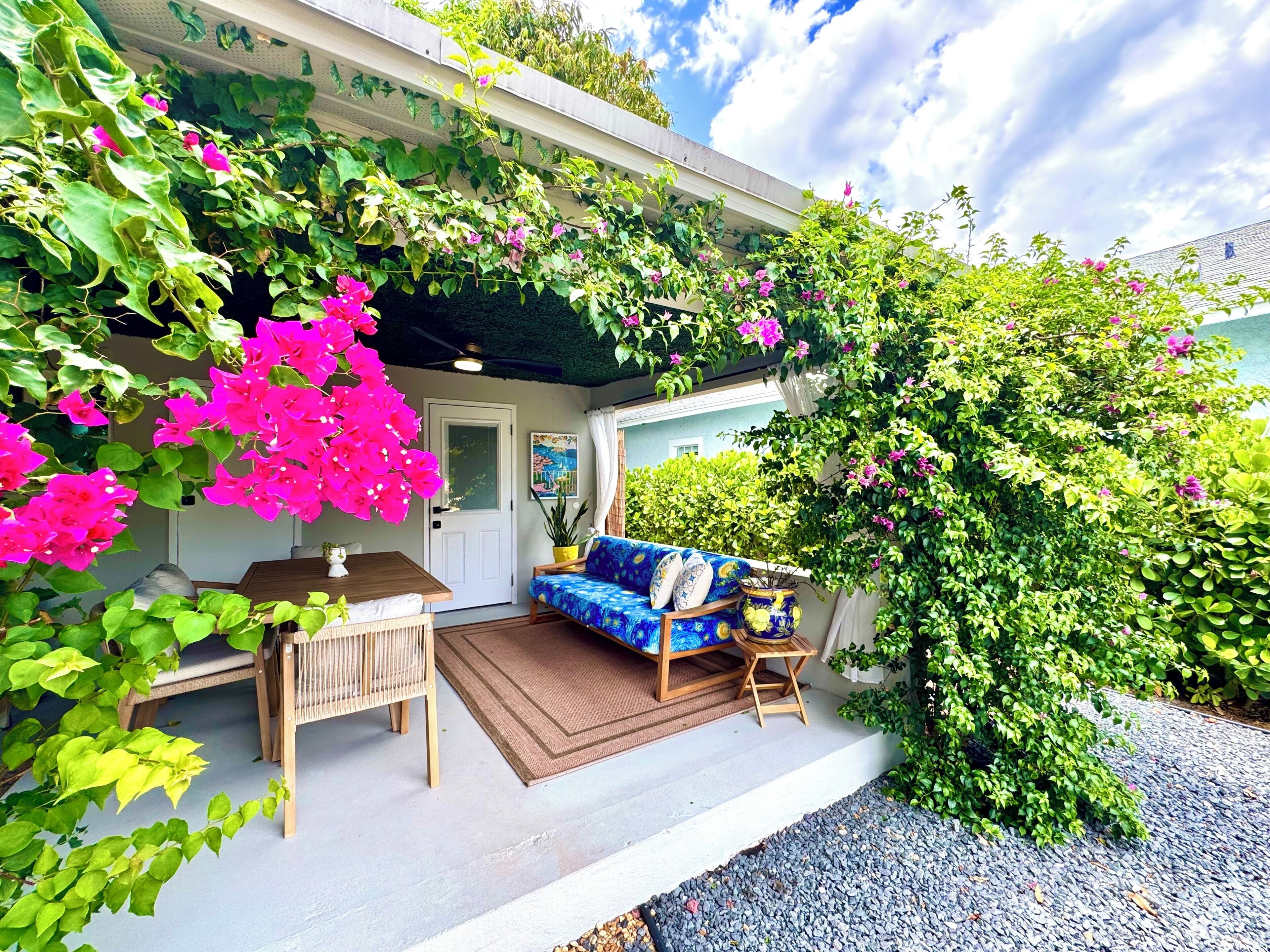 A covered outdoor seating area with a sofa and table, surrounded by lush greenery and flowering plants.