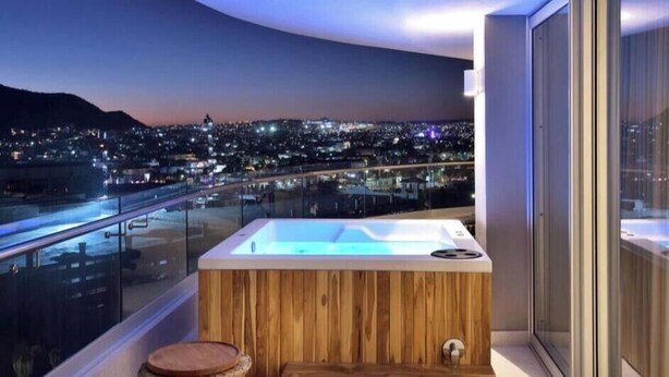 Un balcon doté d’un bain à remous en bois, d’une rambarde en verre et offrant une vue sur la ville la nuit.