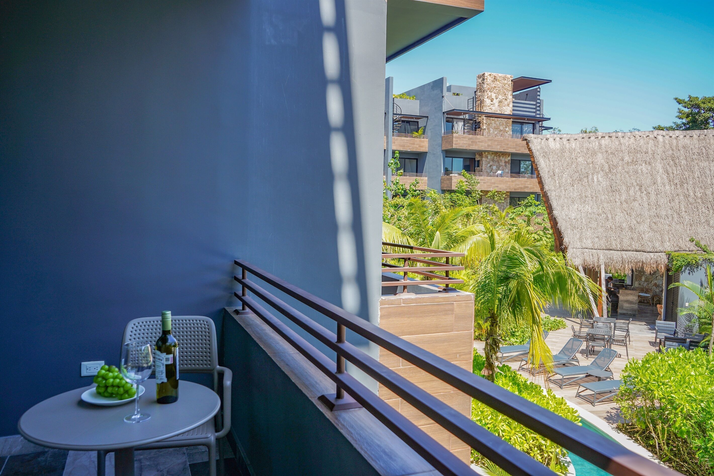 A balcony with a table, a bottle, and a glass of wine, overlooking a pool area with lounge chairs and a building with multiple floors.