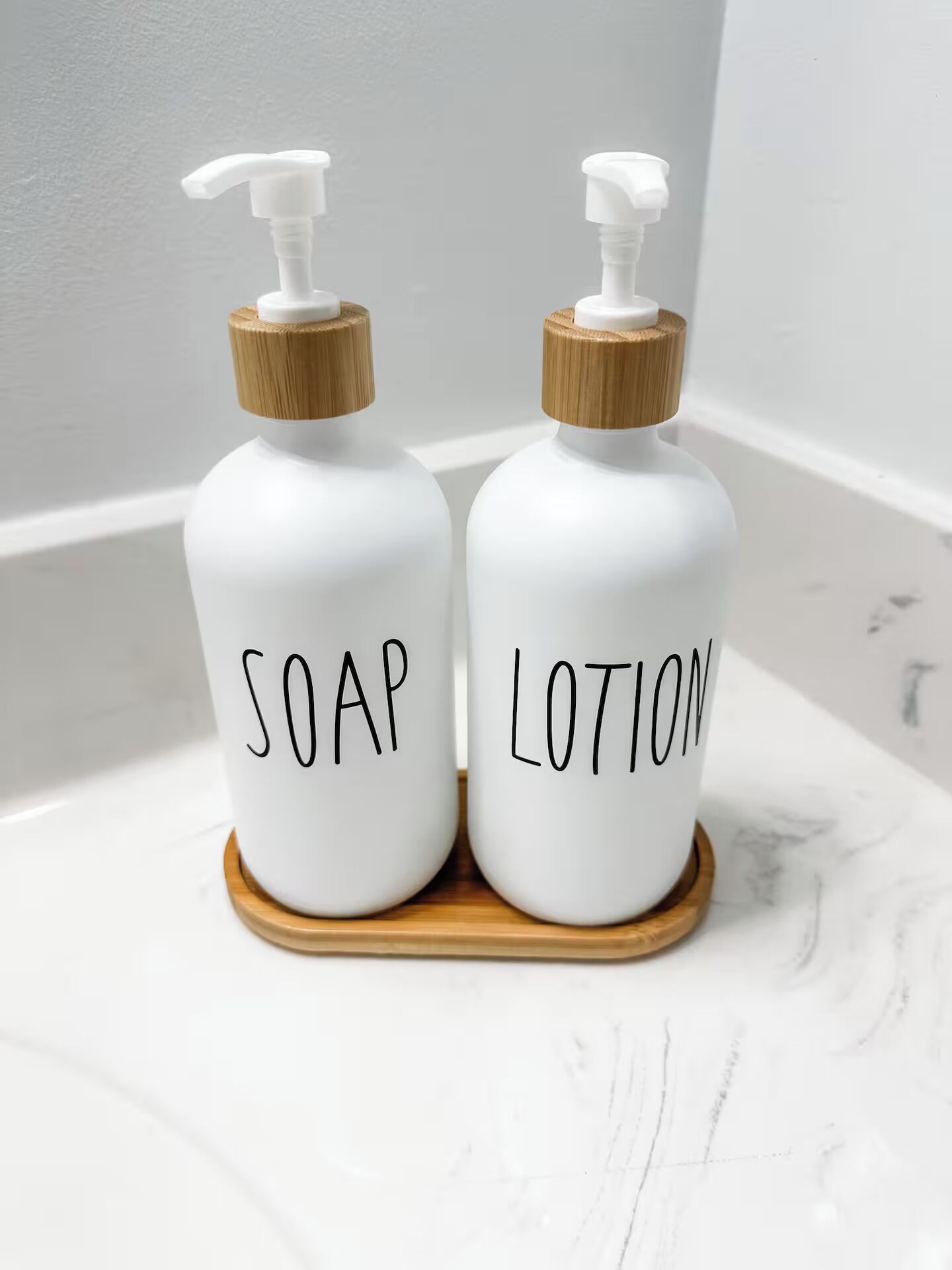 Two white bottles with wooden pumps and labels, one labeled 'SOAP' and the other 'LOTION,' placed on a wooden coaster.
