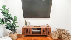 En stue med en træunderholdningsenhed, et fladskærms-TV, en potteplante, en hvid puf og vævede kurve.
