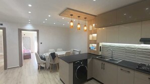 A modern kitchen with a built-in washer, a dining area with a table and chairs, and pendant lighting. - Résidence Oasis (Filfila)