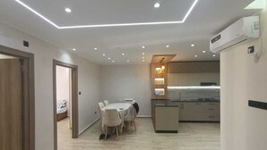 A modern kitchen with a central island, recessed ceiling lighting, and a dining area with a table and chairs. - Résidence Oasis (Filfila)