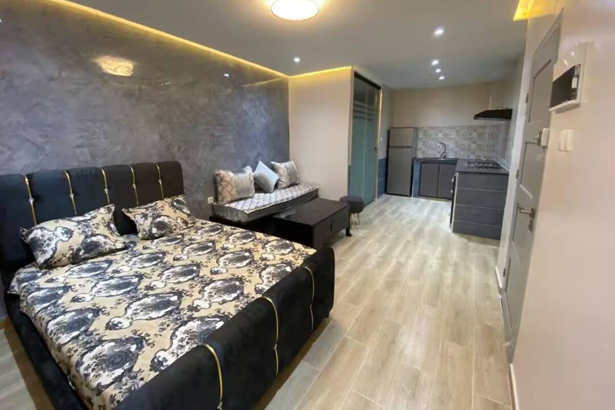 A modern bedroom with a large bed, bedside tables, a built-in bench, and a kitchen area in the background.
