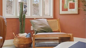Una habitación con una silla de madera, una cesta de mimbre, un cactus y una ventana con vistas a un muro de piedra y una scooter.