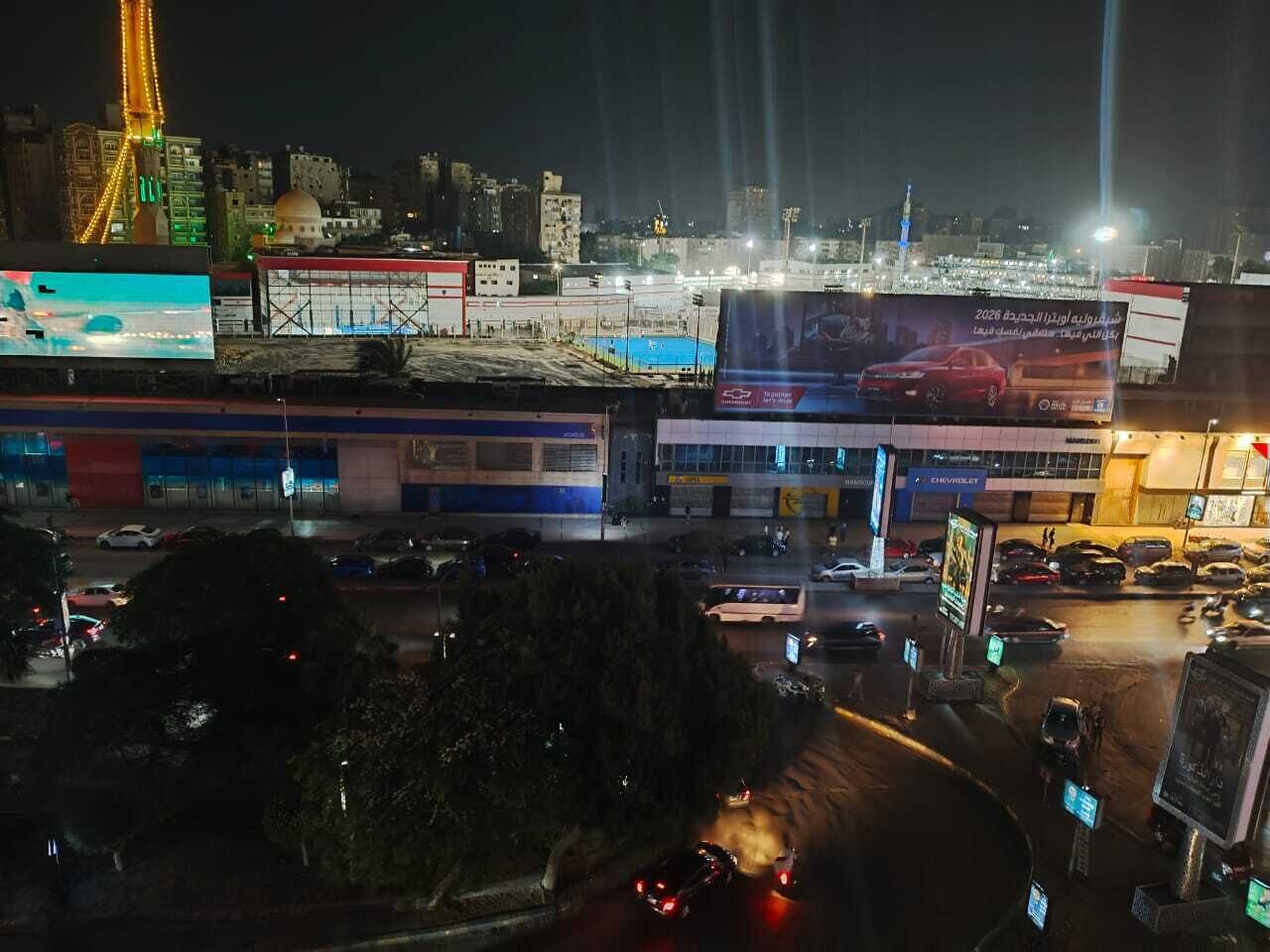 Paisatge urbà nocturn amb edificis il·luminats, una mesquita destacada i un pàrquing concorregut.