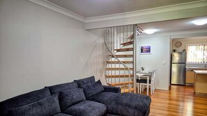 Ein Wohnzimmer mit einer dunkelgrauen Eckcouch, einer Holztreppe, einem Essbereich und einer Küche mit Kühlschrank und Spüle.