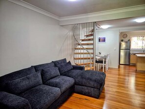 A living room with a dark grey sectional sofa, a wooden staircase, a dining area, and a kitchen with a refrigerator and sink. - Noosaville Getaway, Walk to River, Marina and Boat Ramp (Tewantin)