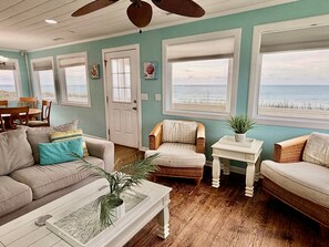 A living room with a view of the ocean, a door, and a dining area with chairs and a table. - Discover 'Diamond in the Rough'—a cozy 3BR oceanfront haven in Oak Island! (Oak Island)