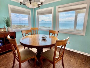 A dining area with a round table, four chairs, a chandelier, and large windows with a view of the ocean. - Discover 'Diamond in the Rough'—a cozy 3BR oceanfront haven in Oak Island! (Oak Island)