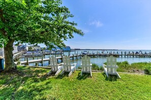 A waterfront area with docked boats, a tree, and three white Adirondack chairs. - 2 Bedroom Bayfront Condo With Pool! (Ocean City)