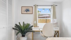 Quarto com cama, escrivaninha com cadeira, planta em vaso, janela com cortinas e quadro na parede.