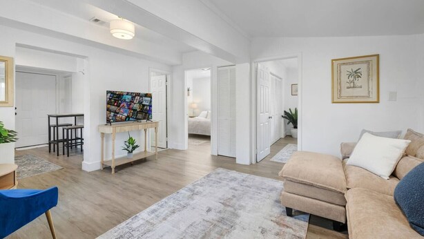 Ampla sala de estar com sofá, televisão em suporte, mesa de jantar e um corredor que dá para um quarto.