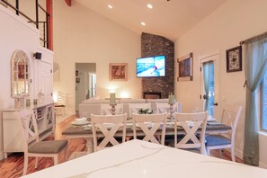 Un comedor con una mesa dispuesta para cuatro, un televisor montado en una pared de piedra, y vista del salón al fondo.