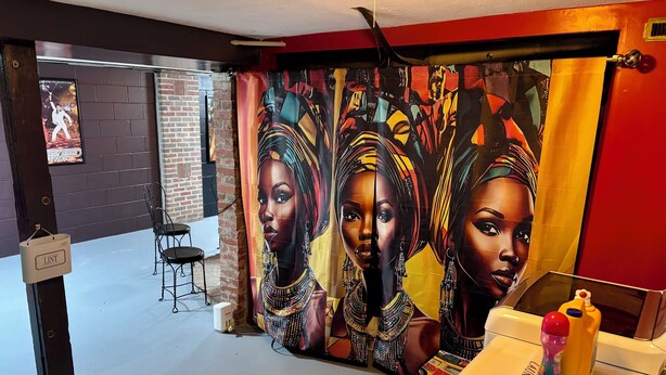A room with a brick wall, a curtain with a colorful design, a small table with a bottle, and a chair.