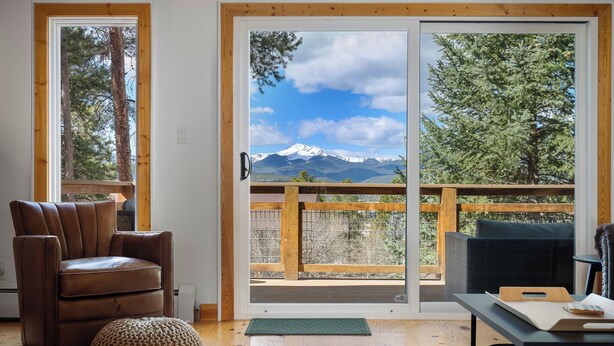 Une pièce comprenant un fauteuil en cuir marron, un pouf rond en fibres tressées, une table en bois avec une planche à découper, et une vue sur une chaîne de montagnes par une porte vitrée.