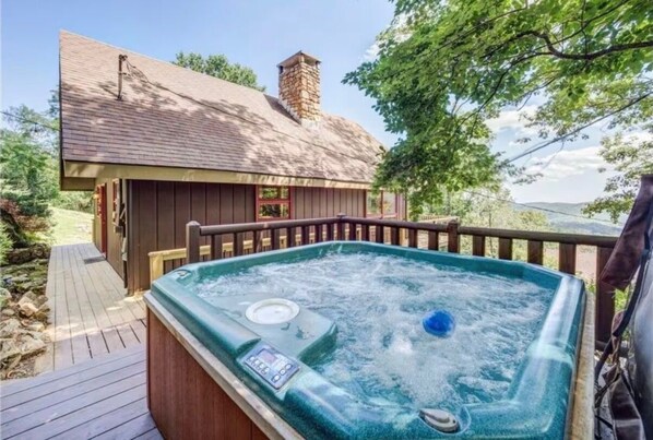 Un bain à remous doté d’un panneau de commande, une terrasse en bois et une maison avec une cheminée en arrière-plan.