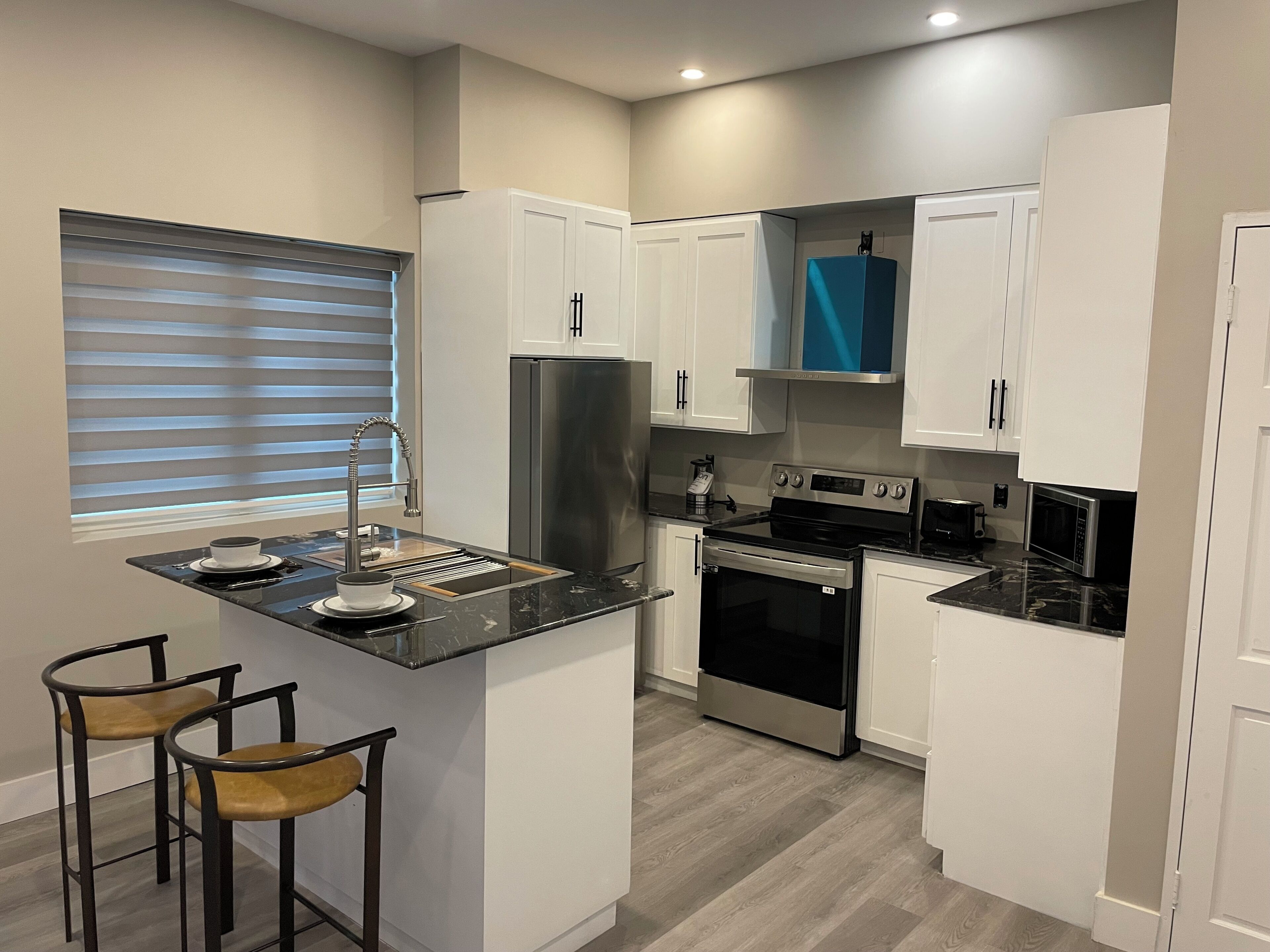 A modern kitchen with white cabinets, a stainless steel refrigerator, a built-in oven, and a microwave. There are two stools and a window with blinds.