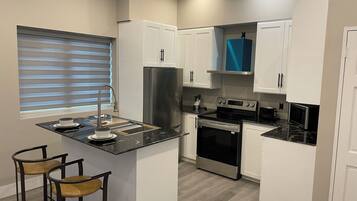 A modern kitchen with white cabinets, a stainless steel refrigerator, a built-in oven, and a microwave. There are two stools and a window with blinds.