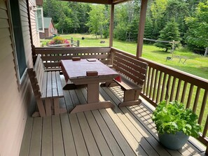 Um deck de madeira com mesa e bancos, cercado por guarda-corpo, com vista para uma área gramada com árvores.