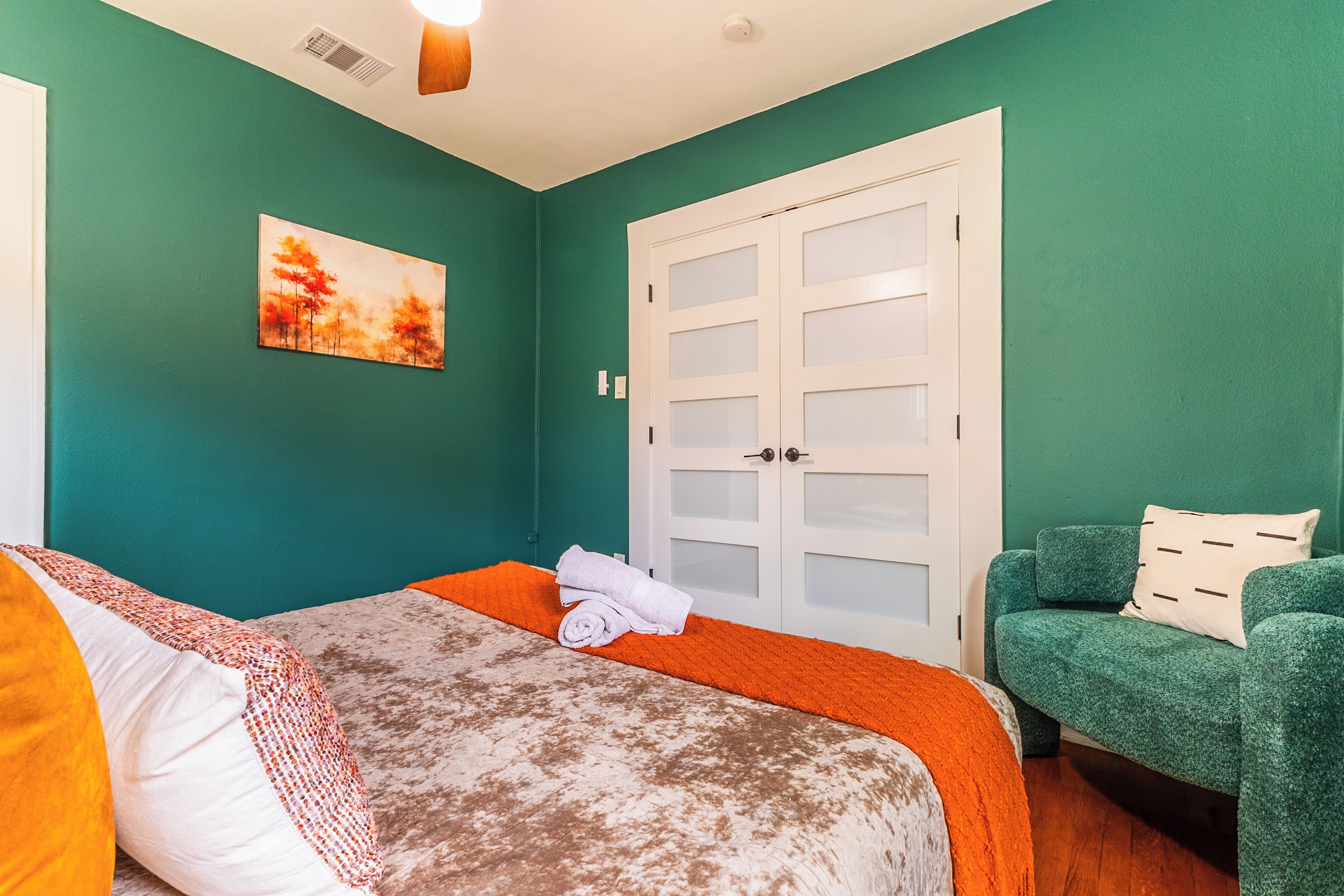 A room with teal walls, a bed with a white and orange bedspread, a green armchair, and a white door with frosted glass panels.