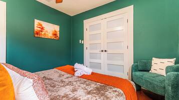 A room with teal walls, a bed with a white and orange bedspread, a green armchair, and a white door with frosted glass panels.