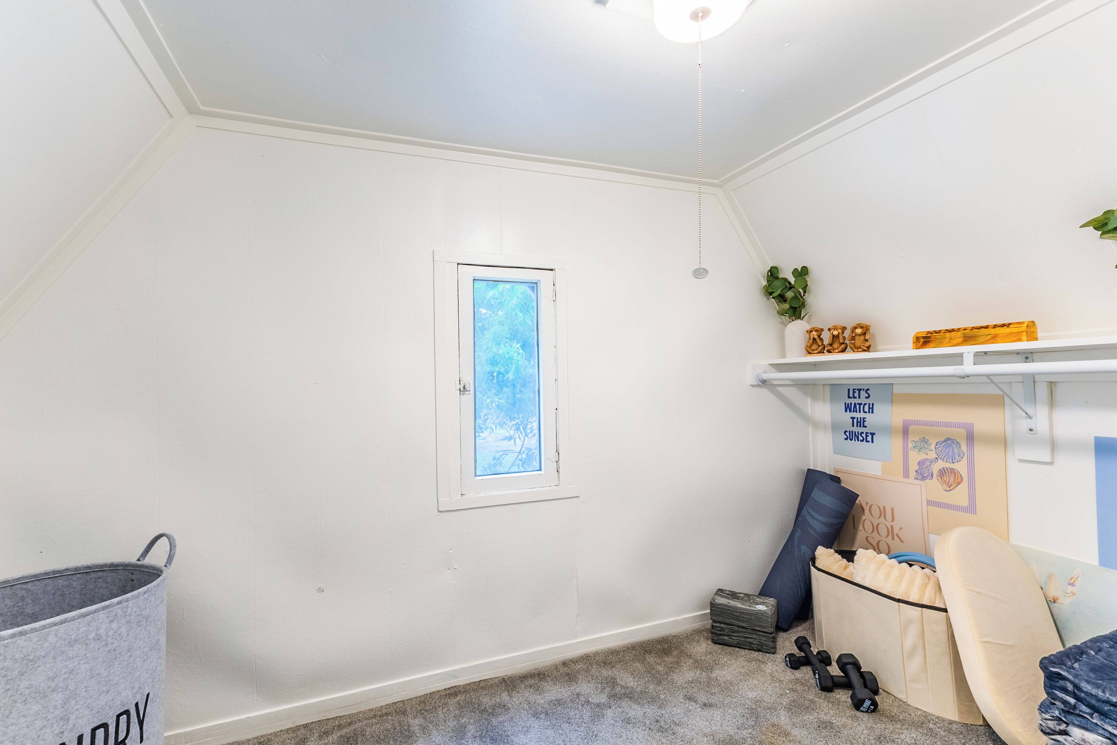 A room with a sloped ceiling, a window with a view, a shelf with decorative items, and a storage unit with exercise equipment.