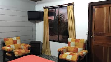 Una habitación con cama, dos sillones, televisión montada en la pared y ventana con cortinas.