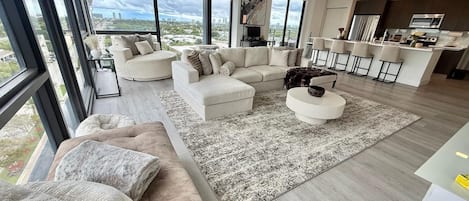 A modern living room with a large window, a sofa set, a coffee table, and a kitchen area with bar stools.