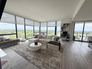 Un salon spacieux avec un grand canapé d’angle, une table basse ronde et une vue sur la ville grâce à de grandes fenêtres.