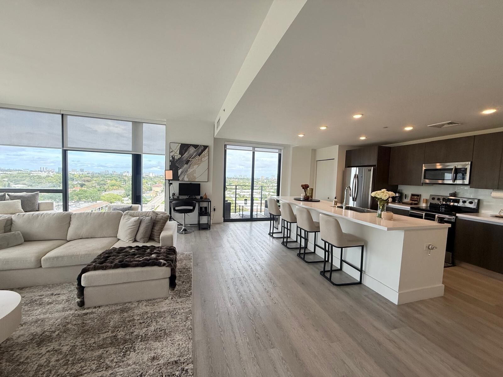 A modern living room with a sofa and ottoman, a kitchen with a bar area, and large windows offering a city view.