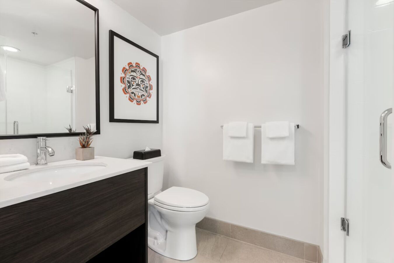 A modern bathroom with a white sink, dark wood vanity, a white toilet, and a framed artwork on the wall.