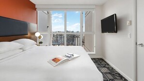Ett hotellrum med en säng, en tv, ett fönster med utsikt över staden och en dörr.