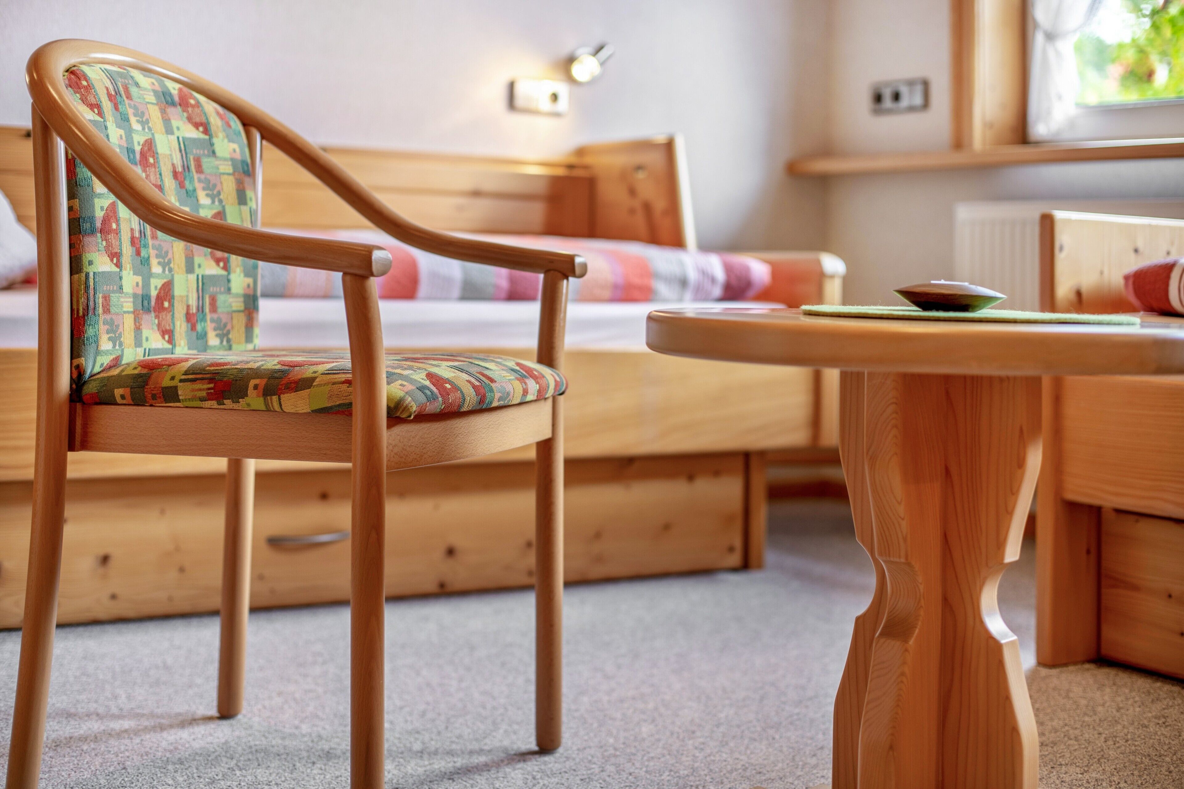 A wooden bed with a headboard, a wooden chair with a colorful cushion, and a small wooden table with a bowl on it.