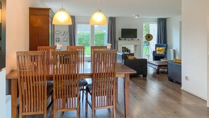 Un comedor con sillas y mesa de madera, una sala de estar con sofá y televisor, y vistas al exterior con zonas verdes.
