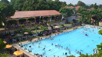 A large swimming pool with people swimming and playing, surrounded by greenery and buildings.
