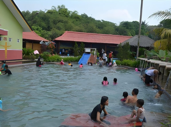 A swimming pool with people in the water, a slide, and a building with a red roof. - Owabong Cottage (Purbalingga)