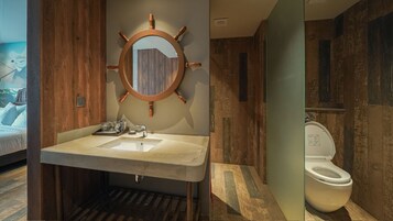 Un baño con tocador de madera, espejo redondo con diseño de timón, inodoro y zona de ducha.