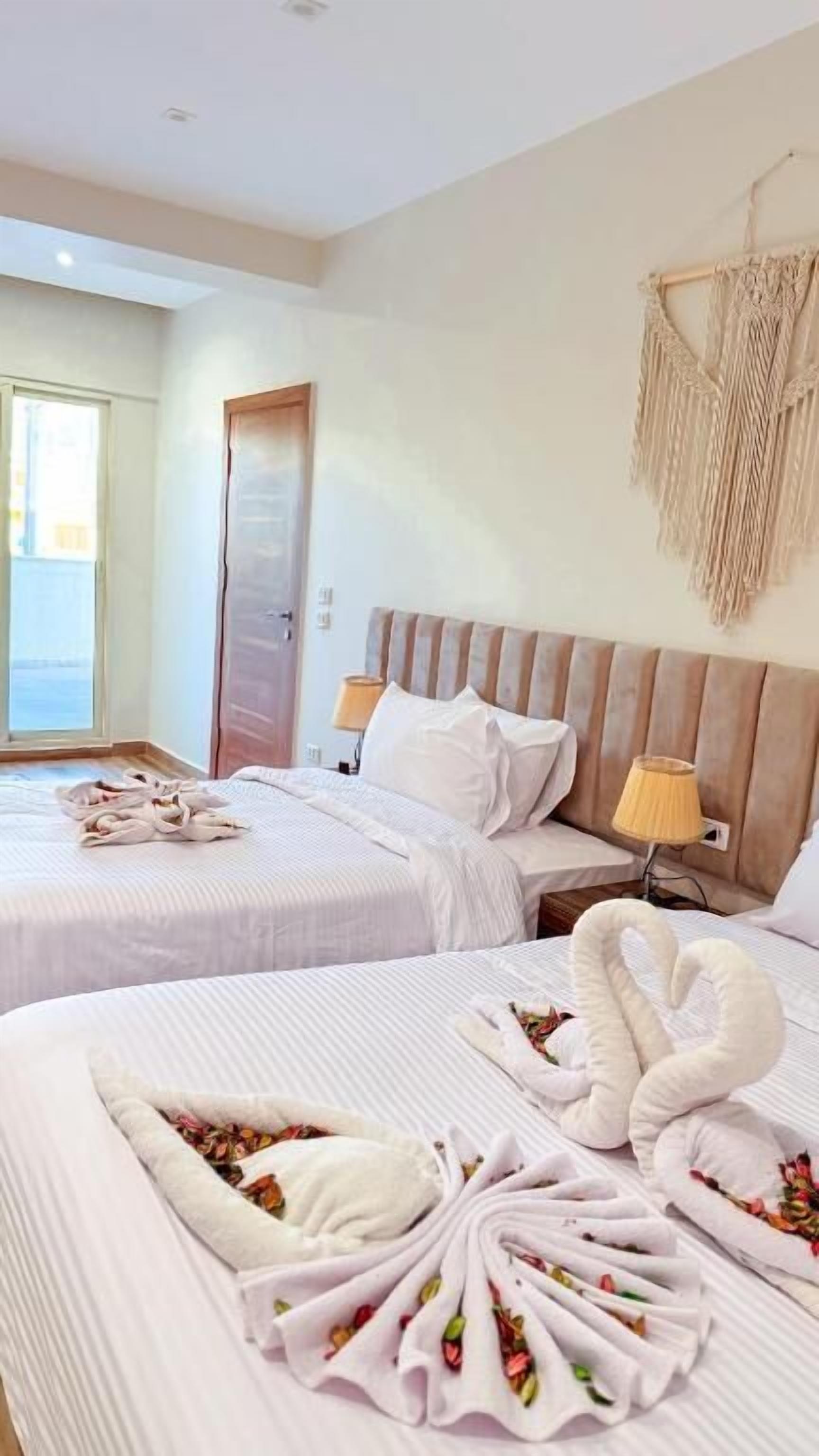A hotel room with two beds, a wooden headboard, a bedside lamp, a macrame wall hanging, and a view through a window.