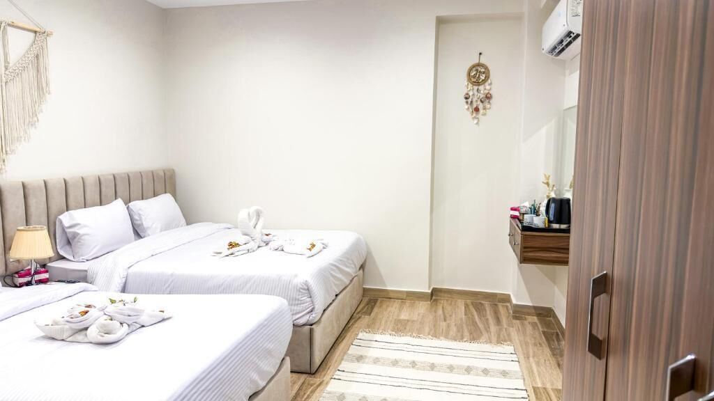 Une chambre d’hôtel avec deux lits, une table de chevet avec une lampe, une petite étagère avec des articles de toilette et un plancher en bois.
