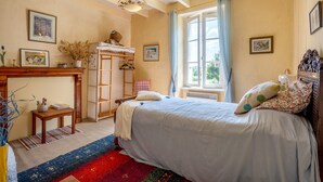 Ein Schlafzimmer mit einem Bett, einer Holzkommode, einem Fenster mit Vorhängen und einem Wandregal.