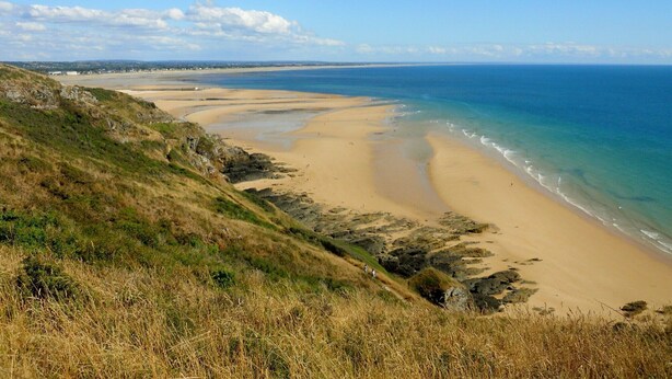 Un paysage côtier avec une plage de sable, des collines verdoyantes et un ciel bleu dégagé.