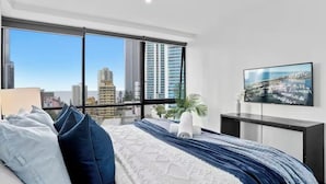 A modern bedroom with a city view, a bed with blue and white bedding, a flat-screen TV, and a small plant. - Modern 2BR Oasis with Pools, Gym and Secure Parking (Surfers Paradise)