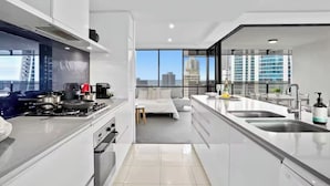 A modern kitchen with a stove, oven, and sink, a dining area with a table and chairs, and large windows offering a city view. - Modern 2BR Oasis with Pools, Gym and Secure Parking (Surfers Paradise)