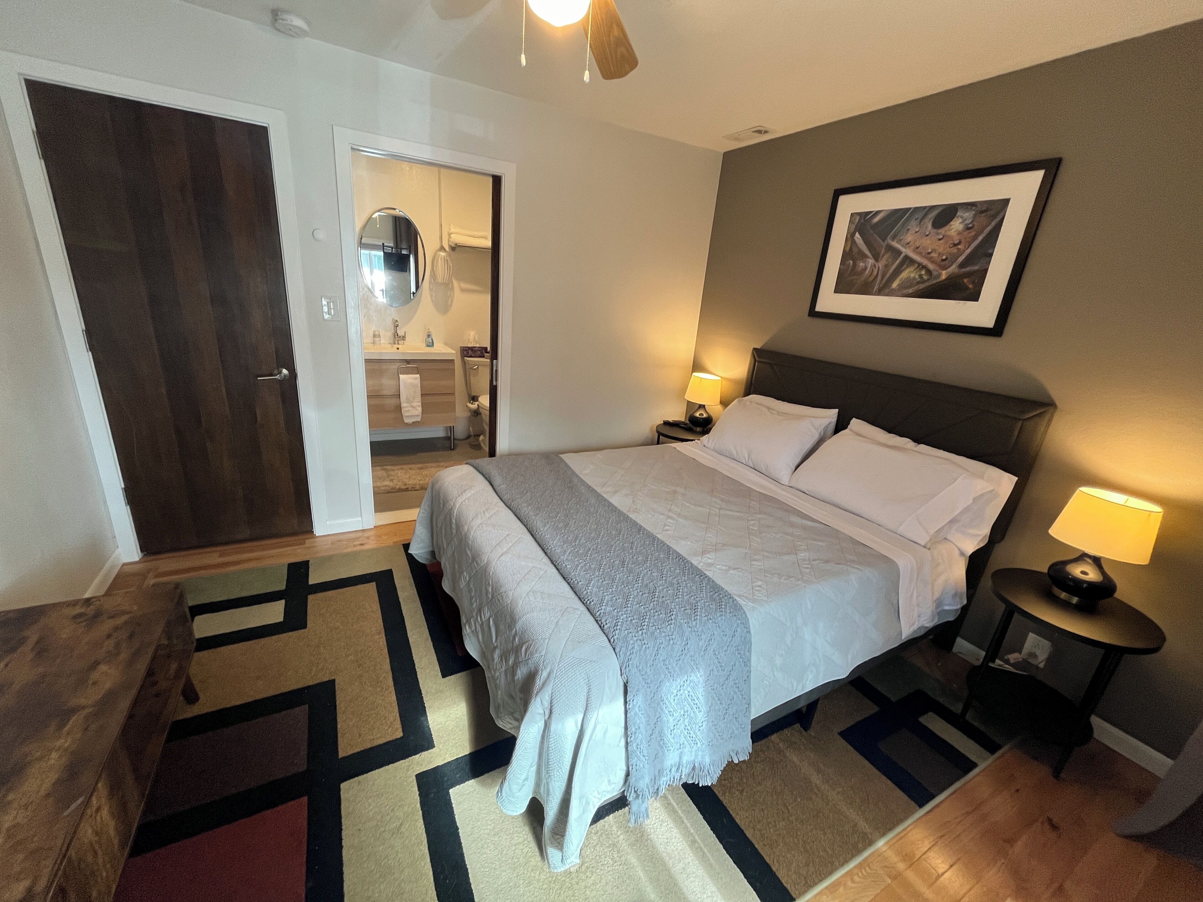 Ein Hotelzimmer mit Bett, Nachttischen, einer Tür, einem sichtbaren Badezimmer durch eine geöffnete Tür und einem gerahmten Bild an der Wand.