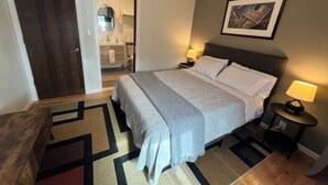 Habitación de hotel con cama, mesitas de noche, un baño visible a través de una puerta entreabierta y un cuadro en la pared.