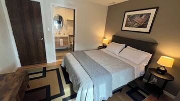Ein Hotelzimmer mit Bett, Nachttischen, einer Tür, einem sichtbaren Badezimmer durch eine geöffnete Tür und einem gerahmten Bild an der Wand.