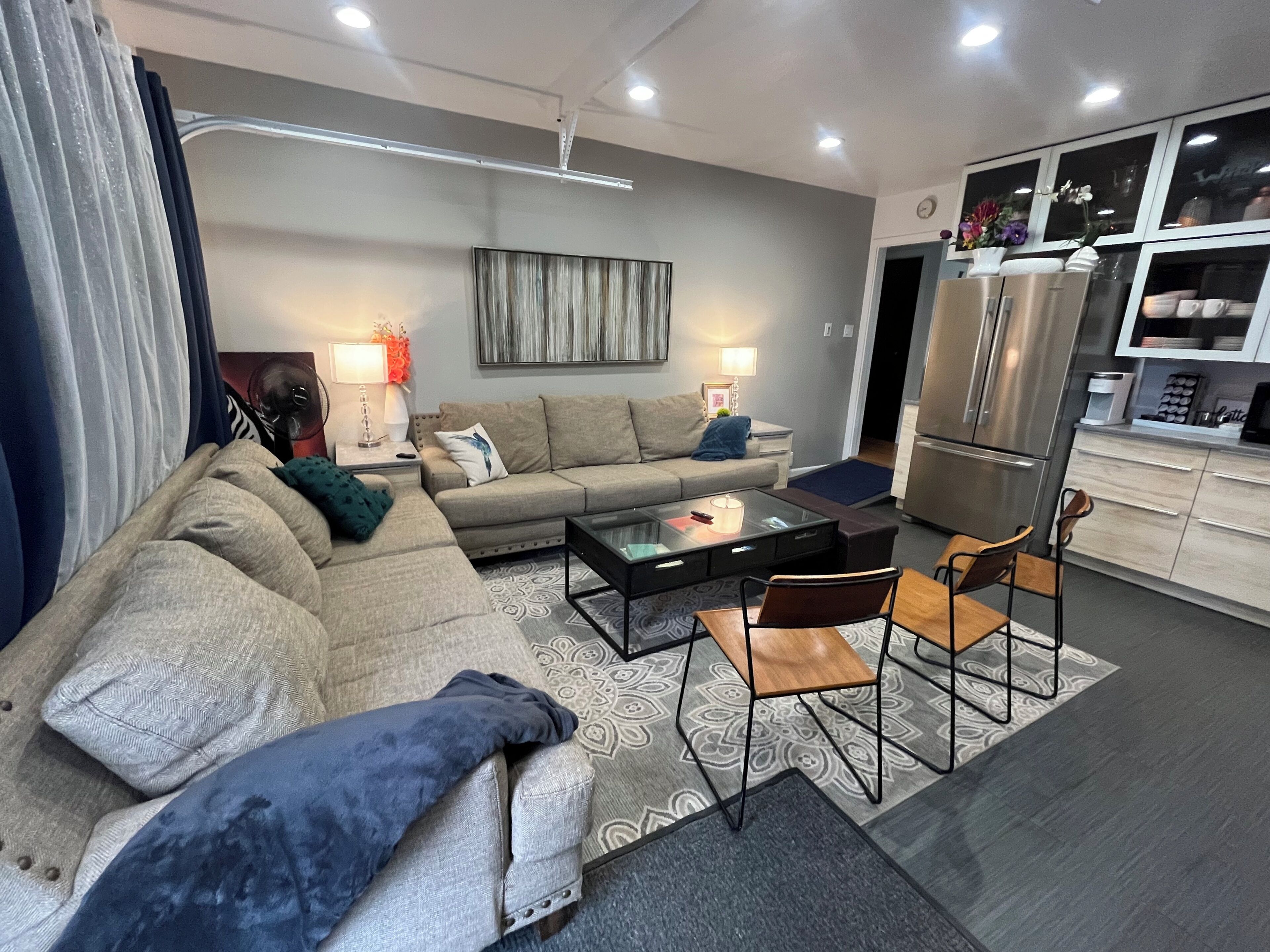 Ein modernes Wohnzimmer mit Sofa, Couchtisch und Sesseln, eine Küchenzeile mit Kühlschrank sowie ein Esstisch.