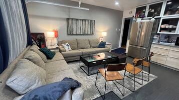Ein modernes Wohnzimmer mit Sofa, Couchtisch und Sesseln, eine Küchenzeile mit Kühlschrank sowie ein Esstisch.